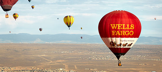 Several hot air balloons in the air against mountainous ground and blue sky. Focus is on the Wells Fargo balloon with logo, stagecoach, and text: Together we'll go far.
